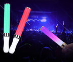 Three glowing foam sticks in green, red, and multicolor held by a person against a concert background.